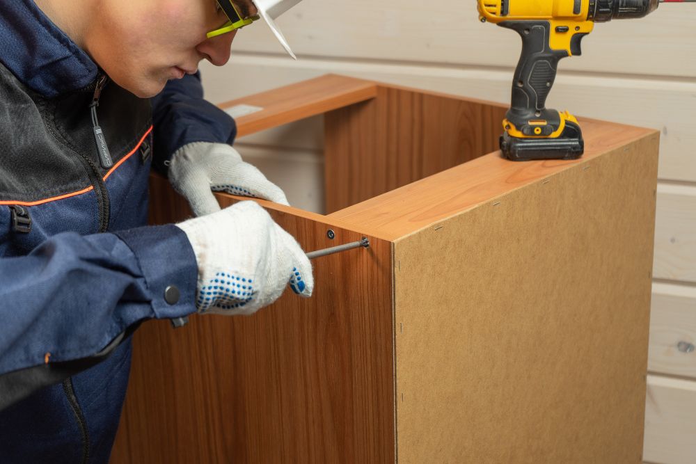 A woman wearing a hat and safety glasses repairs a wooden cabinet with a screwdriver, a drill placed on top.