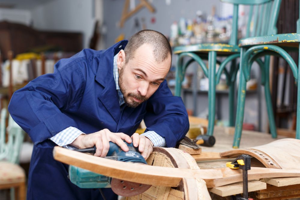 A focused carpenter in a blue shirt sands a wooden chair in a workshop, surrounded by tools and wood shavings.