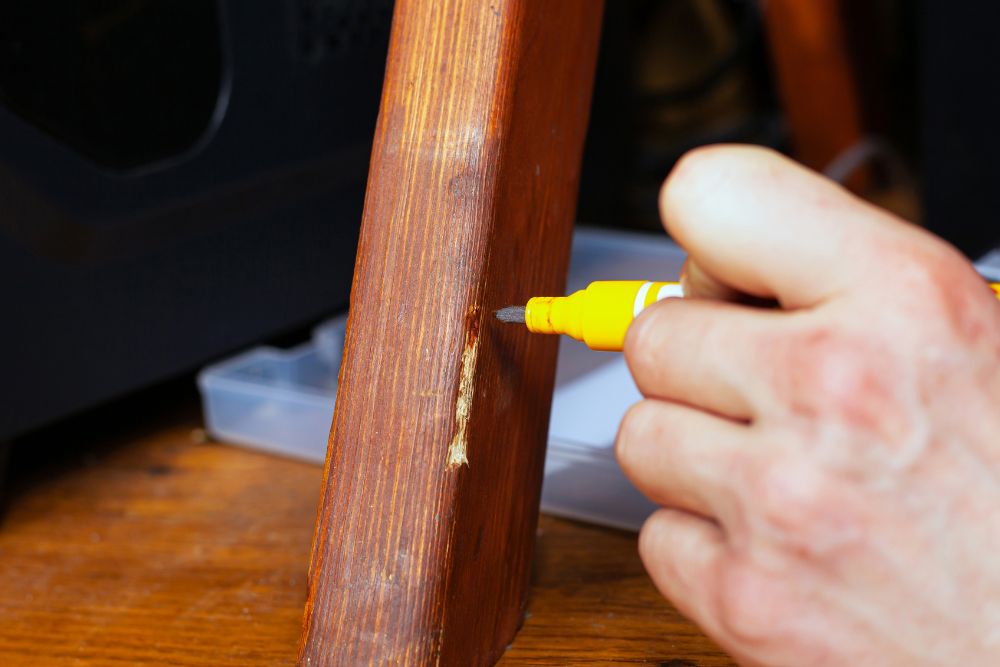 A hand uses a yellow marker to touch up a scratch on a wooden chair leg during a repair process.