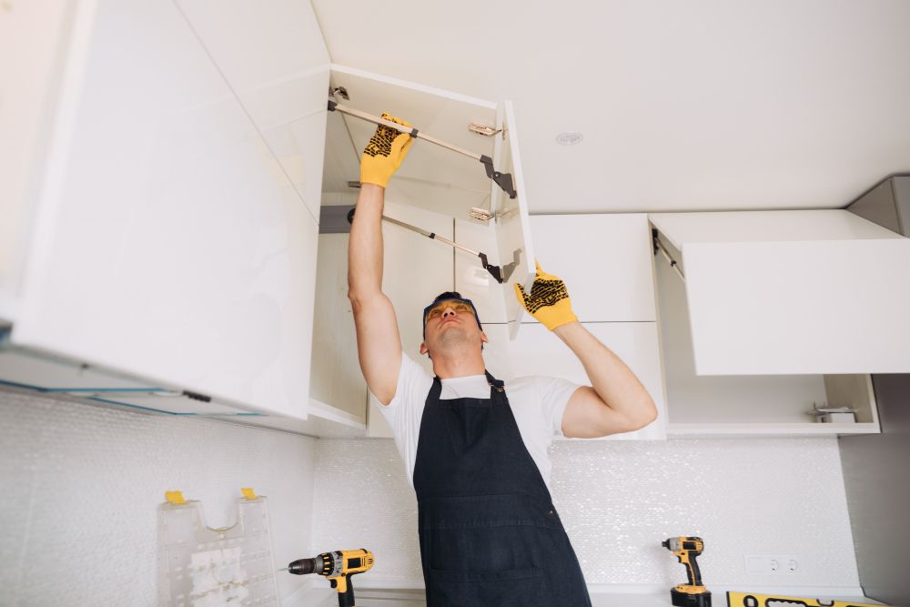 A man wearing an apron repairs a wooden cabinet, focused on his task in a well-lit workshop.