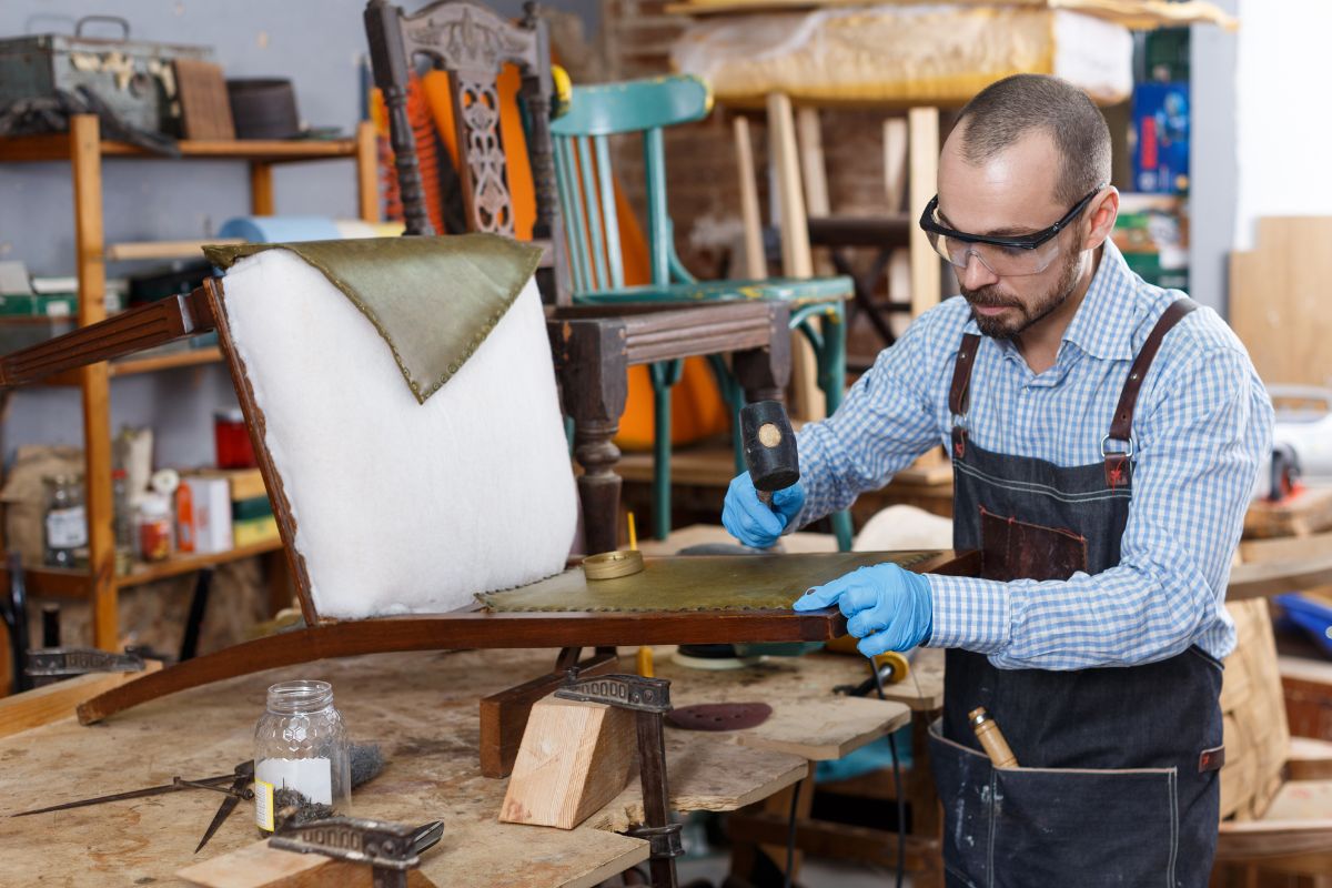  A man wearing an apron is focused on restoring a chair in a furniture service setting.