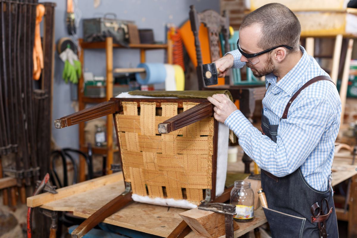 A man repairs a chair in a workshop, surrounded by tools and materials, showcasing furniture service craftsmanship.